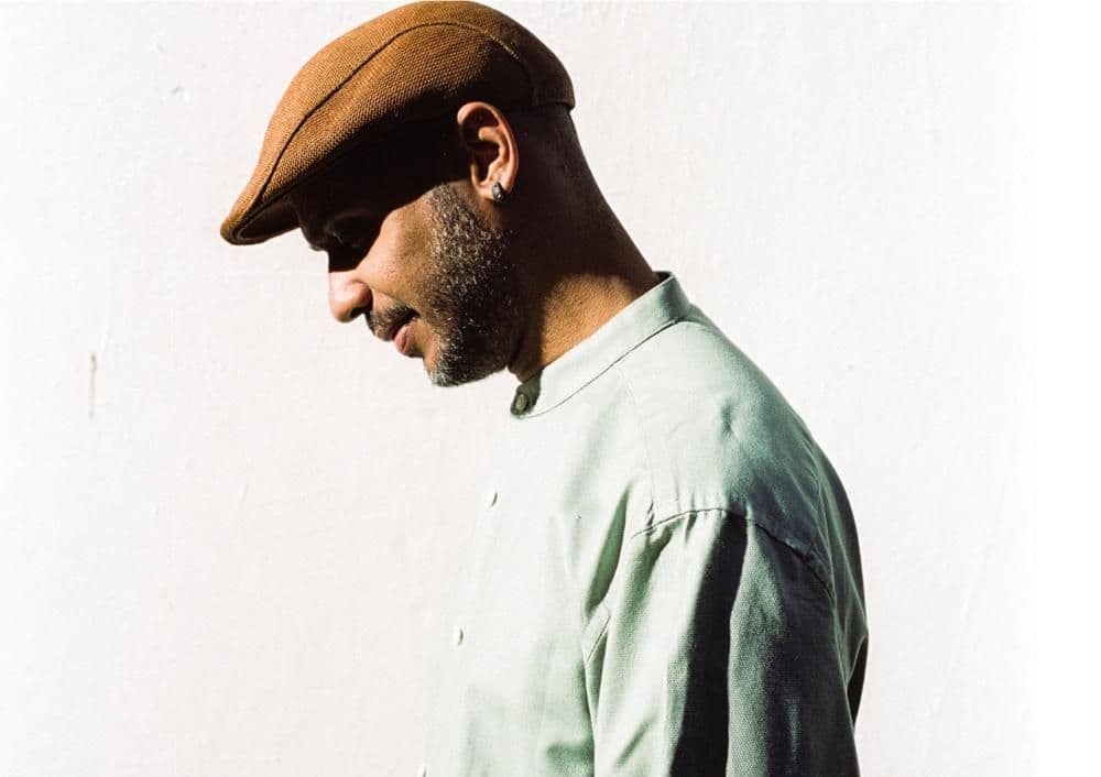A man in a brown hat and green shirt stands, smiling against a neutral background.