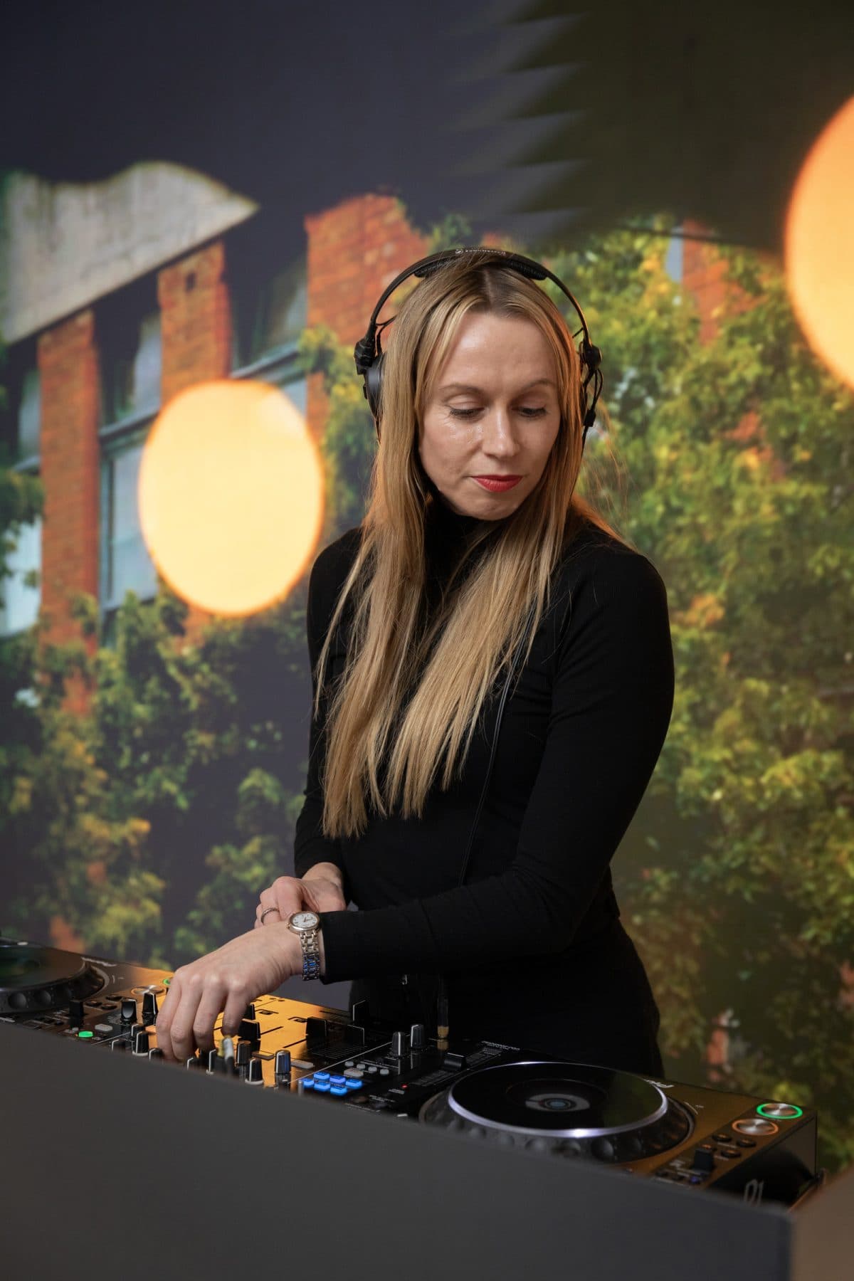 A woman wearing a black shirt and headphones stands at a DJ table, focused on mixing music.